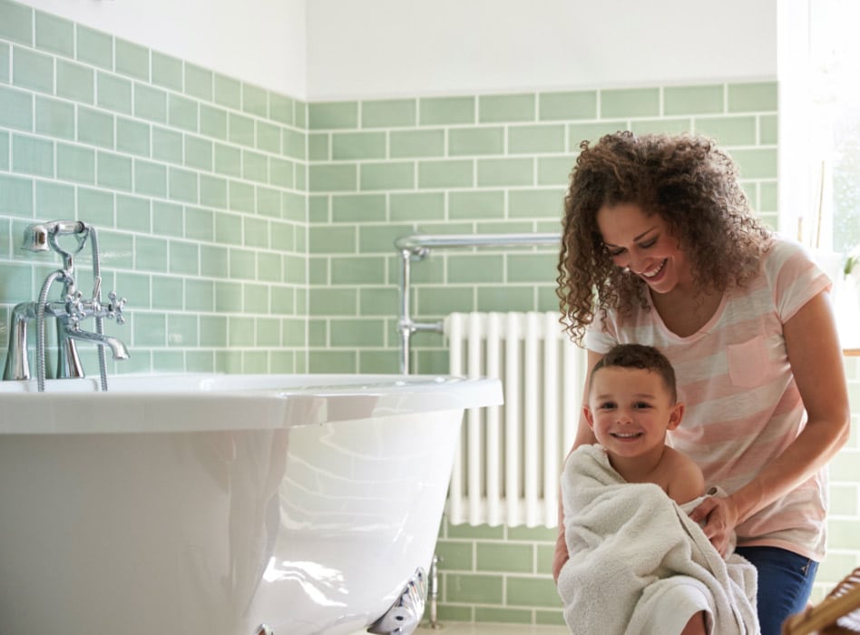 smiling woman drying a young boy wrapped in a towel next to a white bathtub in a bathroom with green tiled walls
