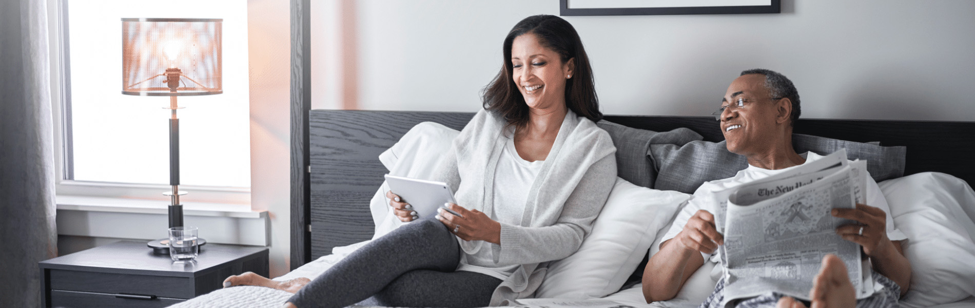 smiling woman sitting on a bed using a tablet while a man beside her reads a newspaper, both relaxed and comfortable in a bright bedroom